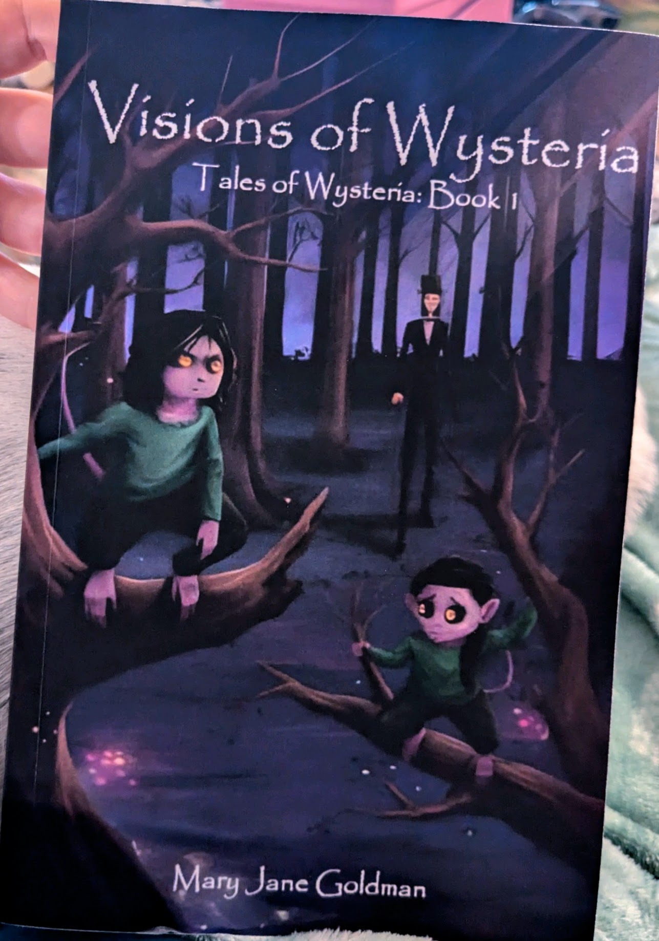 A picture of Visions of Wysteria, held by a reader. Kith and Mara are in the front, crouched in a tree. They have light purple skin, black hair, tails, and golden eyes. In the background lurks a tall dark figure, staring ominously into the forest. 